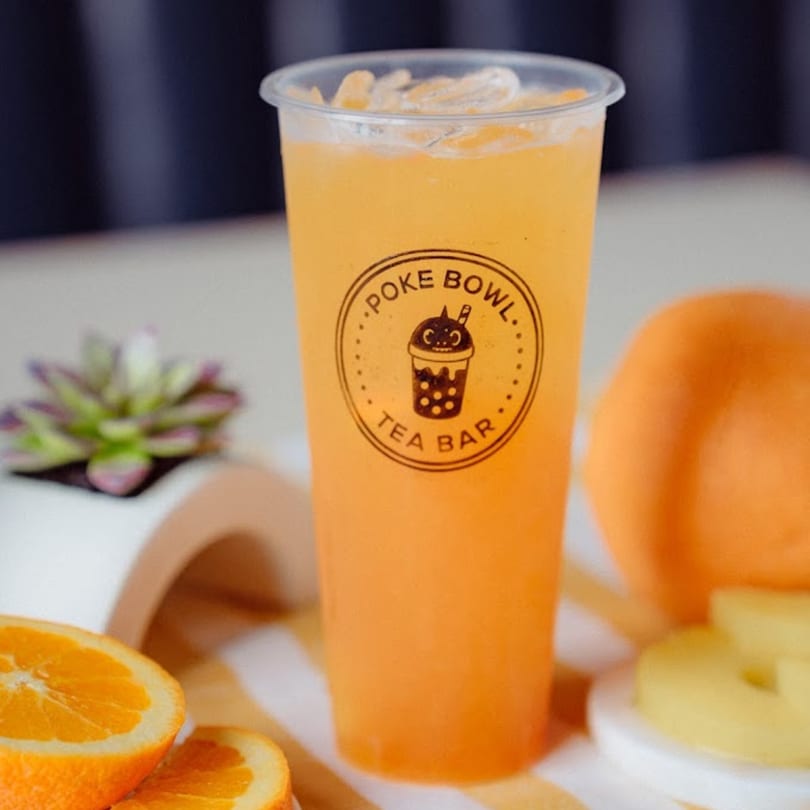A large plastic cup of iced orange tea with the Poke Bowl Tea Bar logo, surrounded by oranges.