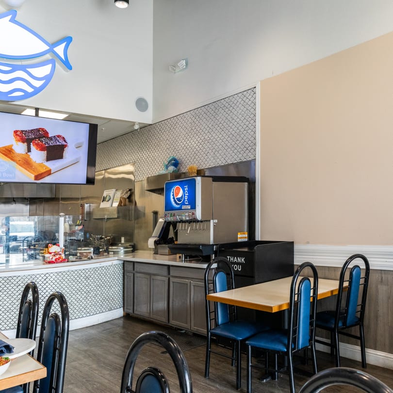Interior of Poke Bowl restaurant with a Pepsi soda fountain, tables, and a sushi display screen.