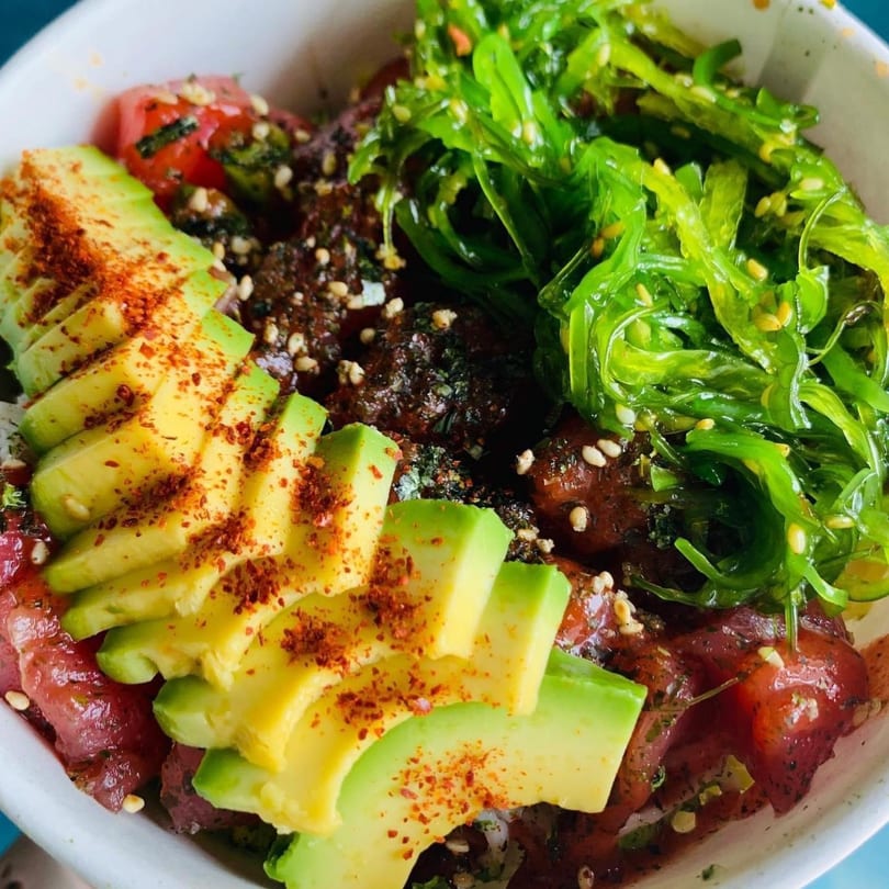 Close-up of a Poke Bowl with tuna, sliced avocado, and bright green seaweed salad.