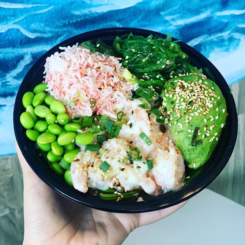 A hand holds a Poke Bowl with shrimp, edamame, avocado, seaweed salad, and imitation crab.
