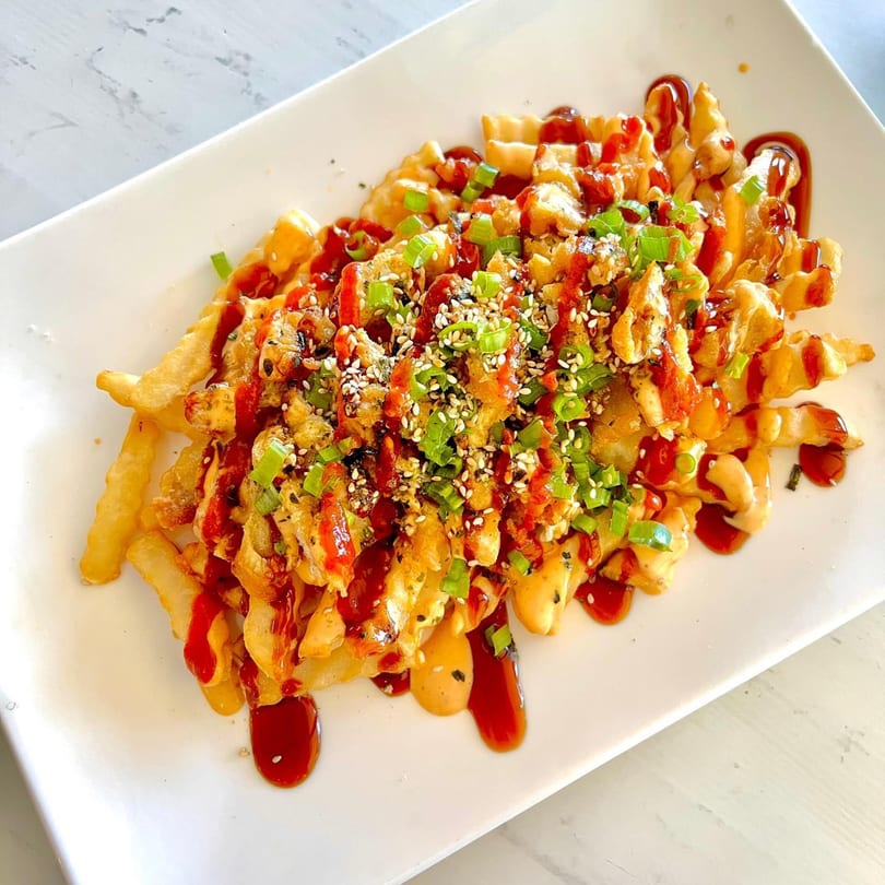 Poke Bowl's loaded fries, topped with sauces, green onions, and sesame seeds on a white plate.