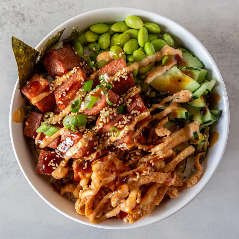 Poke Bowl with spam, edamame, cucumbers, crispy wontons, seaweed, and sauces, garnished with sesame seeds.